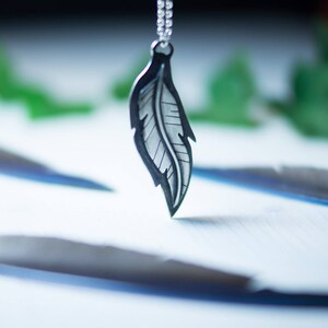 May include: A silver-colored pendant necklace featuring a detailed feather design. The feather has a dark outline and intricate internal lines, suspended from a delicate chain. The background is blurred, with hints of green and white, creating a soft, natural aesthetic.