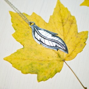May include: A silver-colored feather pendant necklace with a delicate chain. The pendant is detailed with etched lines and rests on a vibrant yellow maple leaf. The leaf's texture and color contrast with the smooth metal, creating a nature-inspired jewelry piece.