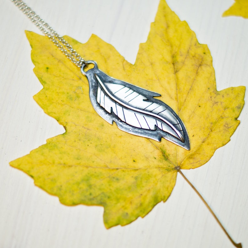 May include: A silver-colored feather pendant necklace with a delicate chain. The pendant is detailed with etched lines and rests on a vibrant yellow maple leaf. The leaf's texture and color contrast with the smooth metal, creating a nature-inspired jewelry piece.