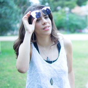May include: A person holding up a pair of sunglasses with a silver frame and blue lenses. The person is wearing a white tank top over a black top, with long silver necklaces. The background is a blurred green park.