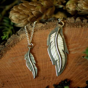 May include: Two silver feather pendants on a wooden surface. One pendant is small and hangs from a chain. The other is larger, with a detailed feather design. The background includes greenery and a brown, textured surface.