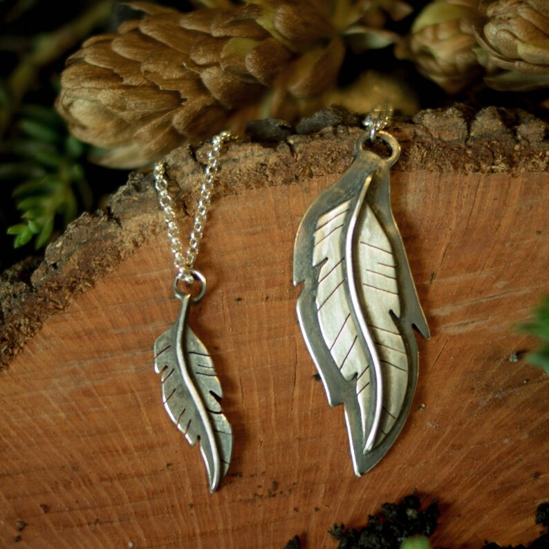 May include: Two silver feather pendants on a wooden surface. One pendant is small and hangs from a chain. The other is larger, with a detailed feather design. The background includes greenery and a brown, textured surface.