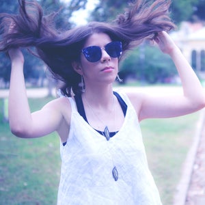 May include: A person wearing blue sequined sunglasses, a white sleeveless top, and a silver necklace with a leaf pendant. The person's hair is styled up. The background is a blurred outdoor scene.