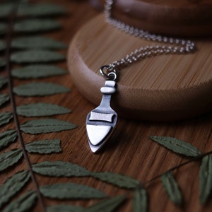 May include: A silver necklace with a pendant shaped like a potion bottle. The pendant has a textured surface and a silver cap. The necklace is on a wooden surface.