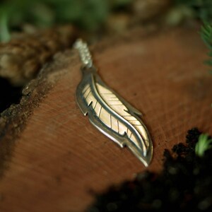 May include: A silver and gold feather pendant necklace. The pendant has a detailed feather design with a silver outline and a gold-colored interior. The necklace is displayed on a wooden surface, with a blurred background of greenery.
