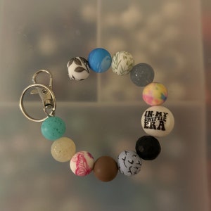 May include: A colourful beaded keychain with a silver key ring. The beads feature various patterns, including floral designs, butterflies, and a bead with the text "In My Swiftie Era".