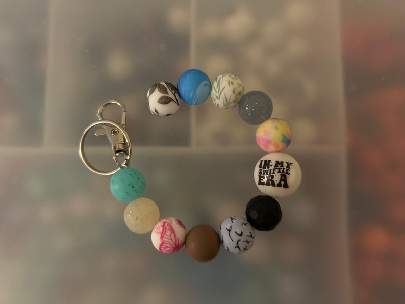 May include: A colourful beaded keychain with a silver key ring. The beads feature various patterns, including floral designs, butterflies, and a bead with the text "In My Swiftie Era".
