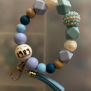 May include: A blue and white beaded keyring with a charm that says "1989 (Taylor's Version)". The keyring has a tassel and a silver clasp.