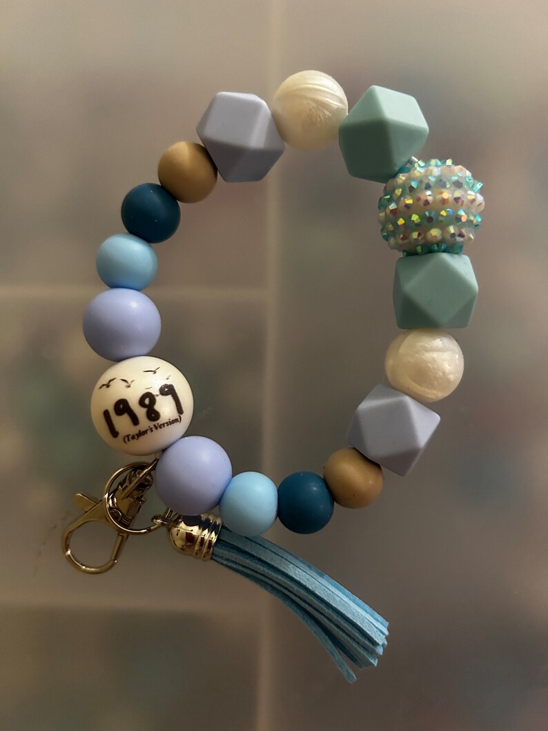 May include: A blue and white beaded keyring with a charm that says "1989 (Taylor's Version)". The keyring has a tassel and a silver clasp.
