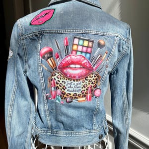 May include: Light blue denim jacket with a makeup-themed graphic on the back. The graphic includes a large pair of red lips, a leopard print makeup bag, and various makeup items. The text "Just a girl who loves makeup" is also visible.