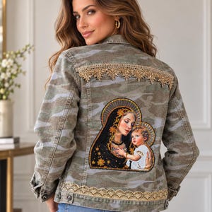 May include: A camouflage print denim jacket featuring a beaded patch of a mother and child on the back. The jacket is embellished with gold lace trim across the upper back and along the bottom hem, with metal buttons on the cuffs and front.