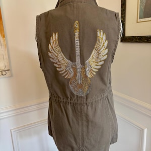 May include: Olive green vest with a hood and a drawstring waist. The back features a sparkling guitar design with wings, embellished with gold and silver rhinestones. The vest is sleeveless and has a casual, yet stylish design.