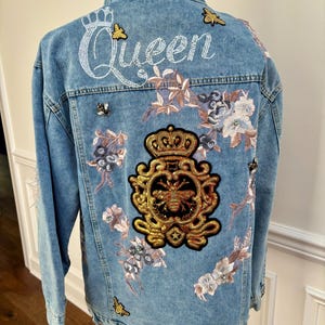 May include: Light blue denim jacket with floral embroidery, butterfly and bee patches. The word "Queen" is embroidered in white script across the upper back. The jacket features a high collar and a distressed look.