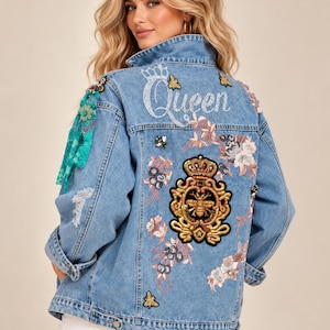 May include: A light blue denim jacket featuring a large embroidered gold bee crest on the back, the word Queen in white script, and various floral and bee patches. The jacket includes turquoise fabric accents on the shoulder and distressed details.