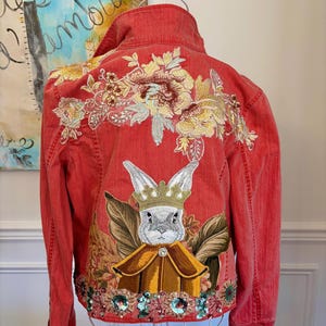 May include: Red denim jacket with elaborate embroidery and sequin embellishments. The back features a regal rabbit design wearing a crown and cape, surrounded by floral patterns. The jacket has a classic collar and button closures.