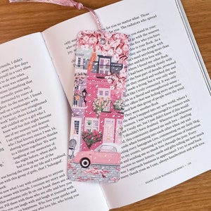 Pode incluir: Um marcador de p&aacute;gina rosa e branco com um carro rosa, um pr&eacute;dio rosa e um design floral. O marcador de p&aacute;gina tem uma borla rosa.