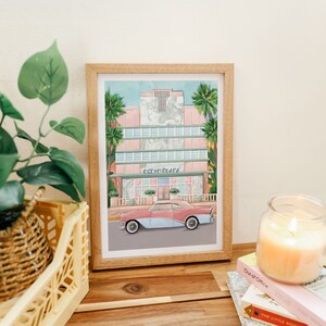 May include: A framed illustration of a pink and white building with the text "Ocean Plaza" above the entrance. A light blue and pink car is parked in front of the building. Palm trees are visible in the background.