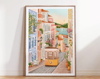 May include: Watercolor print of a European city street scene with a yellow trolley car on cobblestone. Buildings are painted in shades of pink, orange, and blue. The artwork is framed in a light brown wooden frame and leans against a wooden floor.