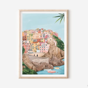 May include: A framed art print depicts a colorful coastal village scene. The buildings are painted in shades of pink, yellow, and orange, set against a light blue sky and turquoise water. Small boats float in the foreground. The print is in a light wood frame.
