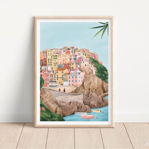 May include: A framed art print depicts a colorful coastal village nestled on a cliffside. The buildings are painted in shades of pink, yellow, and orange. Small boats float in the turquoise water below. The artwork is set against a light blue sky.