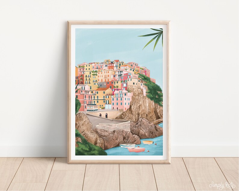 May include: A framed art print depicts a colorful coastal village nestled on a cliffside. The buildings are painted in shades of pink, yellow, and orange. Small boats float in the turquoise water below. The artwork is set against a light blue sky.
