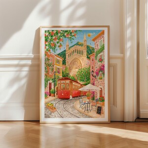 May include: A framed watercolor print depicts a European street scene with a red trolley on tracks. Buildings in shades of orange, pink, and yellow line the cobblestone street. A church and cafe are also visible. The print is in a light wood frame.