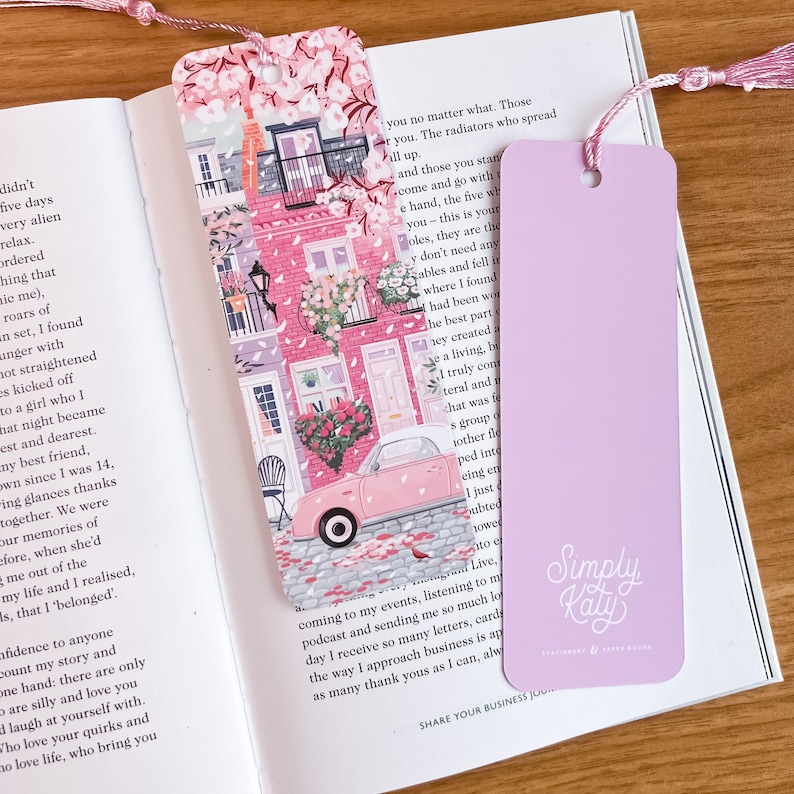 Pode incluir: Um marcador de p&aacute;gina rosa e branco com uma ilustra&ccedil;&atilde;o colorida de um carro rosa dirigindo por uma rua ladeada por edif&iacute;cios rosa. O marcador de p&aacute;gina tem uma borla rosa e o texto "Simply Kaly" no verso.