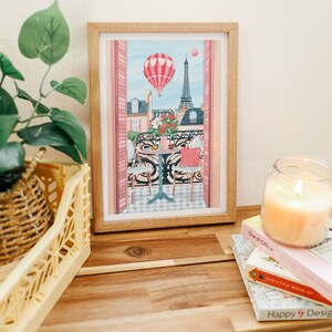 May include: A framed illustration of a Parisian balcony scene with a hot air balloon, Eiffel Tower, and two chairs at a table. The illustration is in a light wood frame and is sitting on a wooden table with books and a candle.