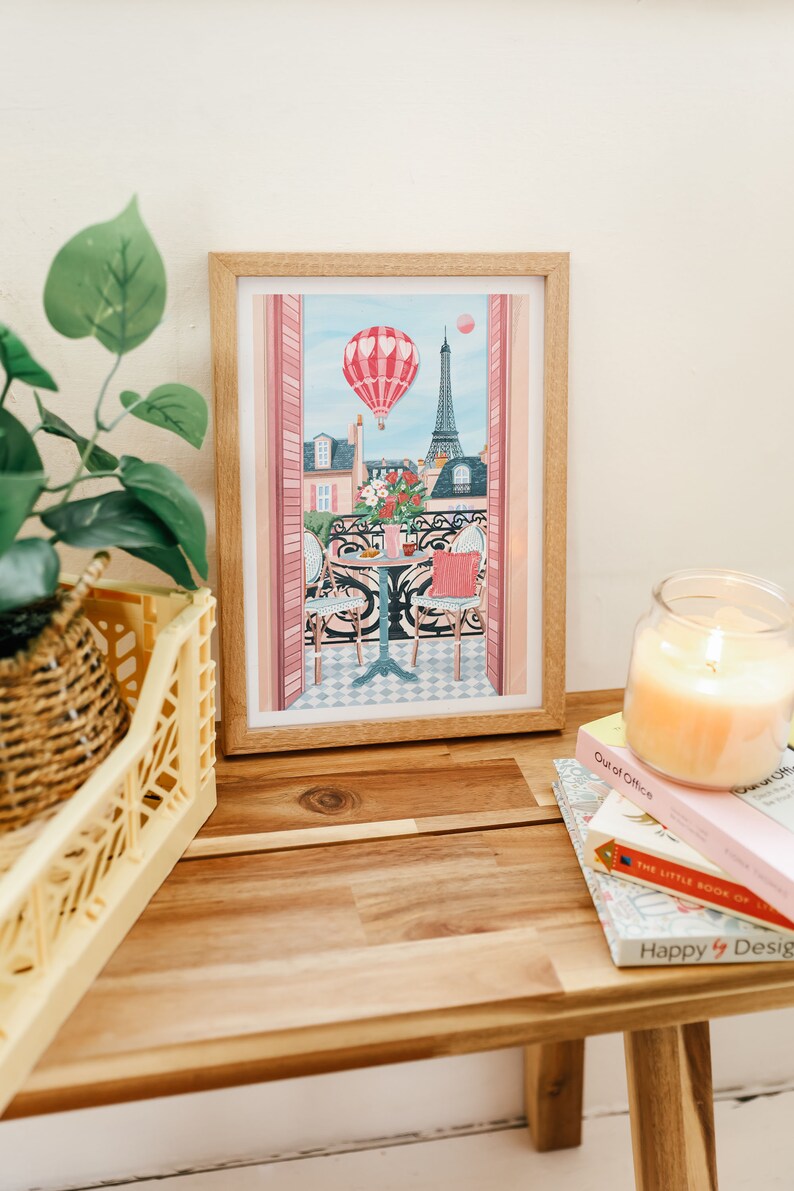 May include: A framed illustration of a Parisian balcony scene with a hot air balloon, Eiffel Tower, and two chairs at a table. The illustration is in a light wood frame and is sitting on a wooden table with books and a candle.