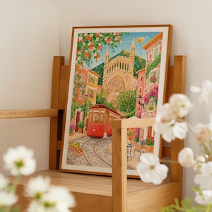 May include: A framed watercolor print depicts a European town scene with a red trolley car on cobblestone tracks. Buildings in shades of pink, orange, and yellow line the street, with a large church in the background. The artwork is displayed on a wooden shelf.