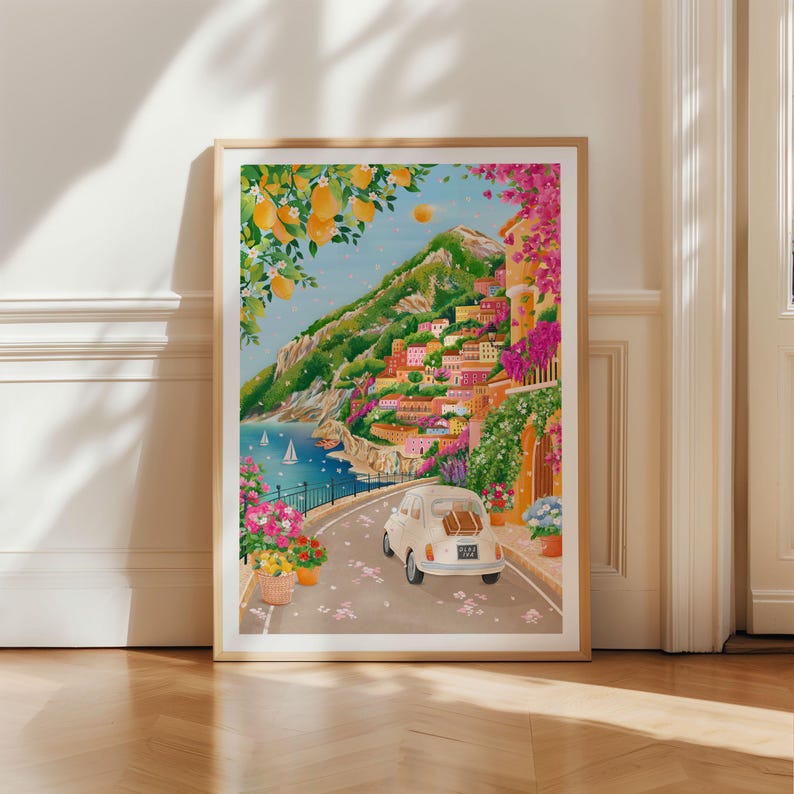 May include: A framed art print depicts a coastal Italian town with a winding road. A vintage white car with luggage drives along the road. The scene includes colorful buildings, lush greenery, and a blue sea with sailboats. Lemon trees frame the top.