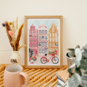 Puede incluir: Una ilustraci&oacute;n enmarcada de una fila de edificios coloridos con una bicicleta roja en frente. Los edificios tienen ventanas blancas y est&aacute;n decorados con flores. La ilustraci&oacute;n est&aacute; sobre un fondo blanco.