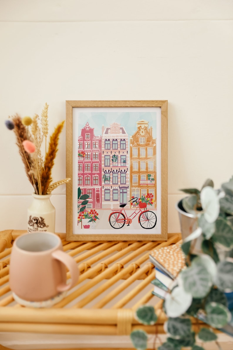 Puede incluir: Una ilustraci&oacute;n enmarcada de una fila de edificios coloridos con una bicicleta roja en frente. Los edificios tienen ventanas blancas y est&aacute;n decorados con flores. La ilustraci&oacute;n est&aacute; sobre un fondo blanco.
