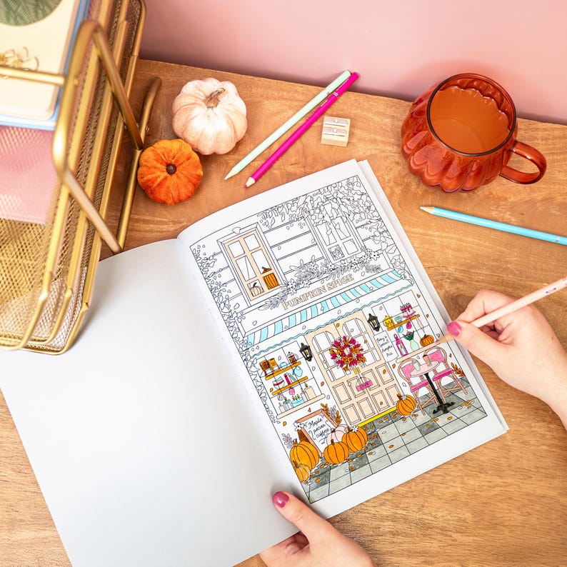 May include: A coloring book page featuring a detailed illustration of a cafe with the text "Pumpkin Spice". The image includes pumpkins, a wreath, and a pink table. Colored pencils and a mug of tea are also present.