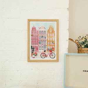 Puede incluir: Una impresi&oacute;n enmarcada de una ilustraci&oacute;n colorida de una fila de casas con una bicicleta en frente. Las casas est&aacute;n pintadas en tonos de rosa, amarillo y azul. La bicicleta es roja con una cesta de flores.