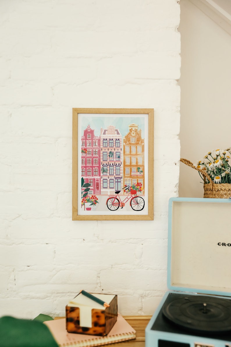 Puede incluir: Una impresi&oacute;n enmarcada de una ilustraci&oacute;n colorida de una fila de casas con una bicicleta en frente. Las casas est&aacute;n pintadas en tonos de rosa, amarillo y azul. La bicicleta es roja con una cesta de flores.