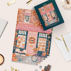 May include: A 500-piece jigsaw puzzle titled Pumpkin Spice Bakery, featuring an illustration of a pink storefront with a striped awning, a bicycle with a flower basket, and autumn foliage. The box displays the same bakery scene.
