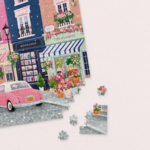 May include: A partially assembled jigsaw puzzle depicting a street scene with a bookshop, florist, and a pink car. The puzzle pieces show buildings with balconies, flowers, and the text "BOOKSHOP" and "THE FLORIST".