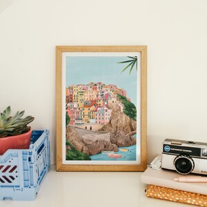 May include: A framed art print depicting a colorful coastal village scene. The artwork features buildings in various colors, a road, boats, and a light blue sky. The print is in a light wood frame, displayed on a white shelf with a blue crate and a vintage camera.