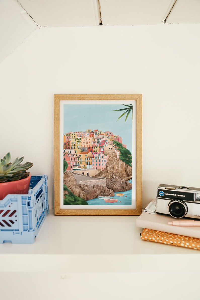 May include: A framed art print depicting a colorful coastal village scene. The artwork features buildings in various colors, a road, boats, and a light blue sky. The print is in a light wood frame, displayed on a white shelf with a blue crate and a vintage camera.