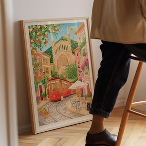 May include: A framed watercolor print depicts a European town scene with a red trolley car on cobblestone tracks. The artwork features a church, buildings, and a cafe with a white umbrella. The print is in a light wood frame.