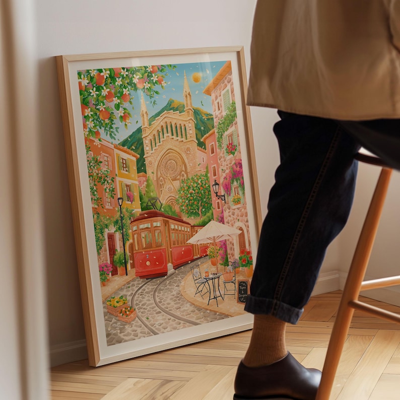 May include: A framed watercolor print depicts a European town scene with a red trolley car on cobblestone tracks. The artwork features a church, buildings, and a cafe with a white umbrella. The print is in a light wood frame.
