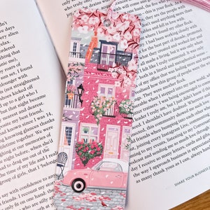 Pode incluir: Marca-p&aacute;ginas rosa e branco com um carro rosa, flores e um pr&eacute;dio rosa com telhado branco. O marca-p&aacute;ginas tem o texto "to".