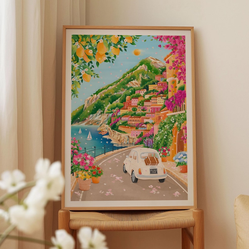 May include: A framed art print depicting a coastal Italian town with colorful buildings nestled on a hillside. A white car drives along a road, and the scene is framed by lemon trees and vibrant flowers. Sailboats dot the blue sea.