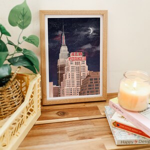 May include: A framed illustration of a tall building with the words "NEW YORKER" in red letters on the side. The building is set against a dark blue night sky with stars and a crescent moon.