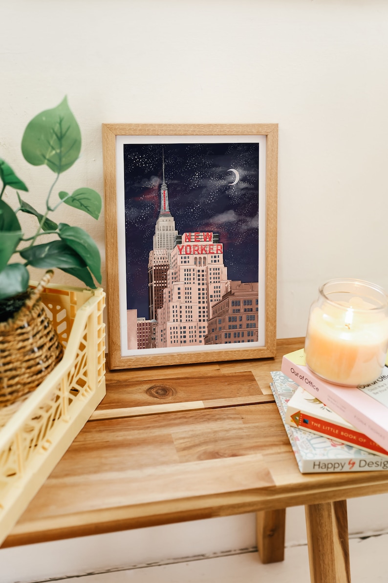 May include: A framed illustration of a tall building with the words "NEW YORKER" in red letters on the side. The building is set against a dark blue night sky with stars and a crescent moon.