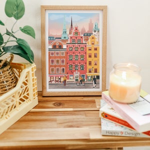 Pode incluir: Uma ilustra&ccedil;&atilde;o emoldurada de uma cena de rua da cidade com edif&iacute;cios coloridos, uma bicicleta e pessoas caminhando. Os edif&iacute;cios est&atilde;o em uma paleta de cores pastel com detalhes em rosa, amarelo e azul. A ilustra&ccedil;&atilde;o &eacute; emoldurada em uma moldura de madeira clara.