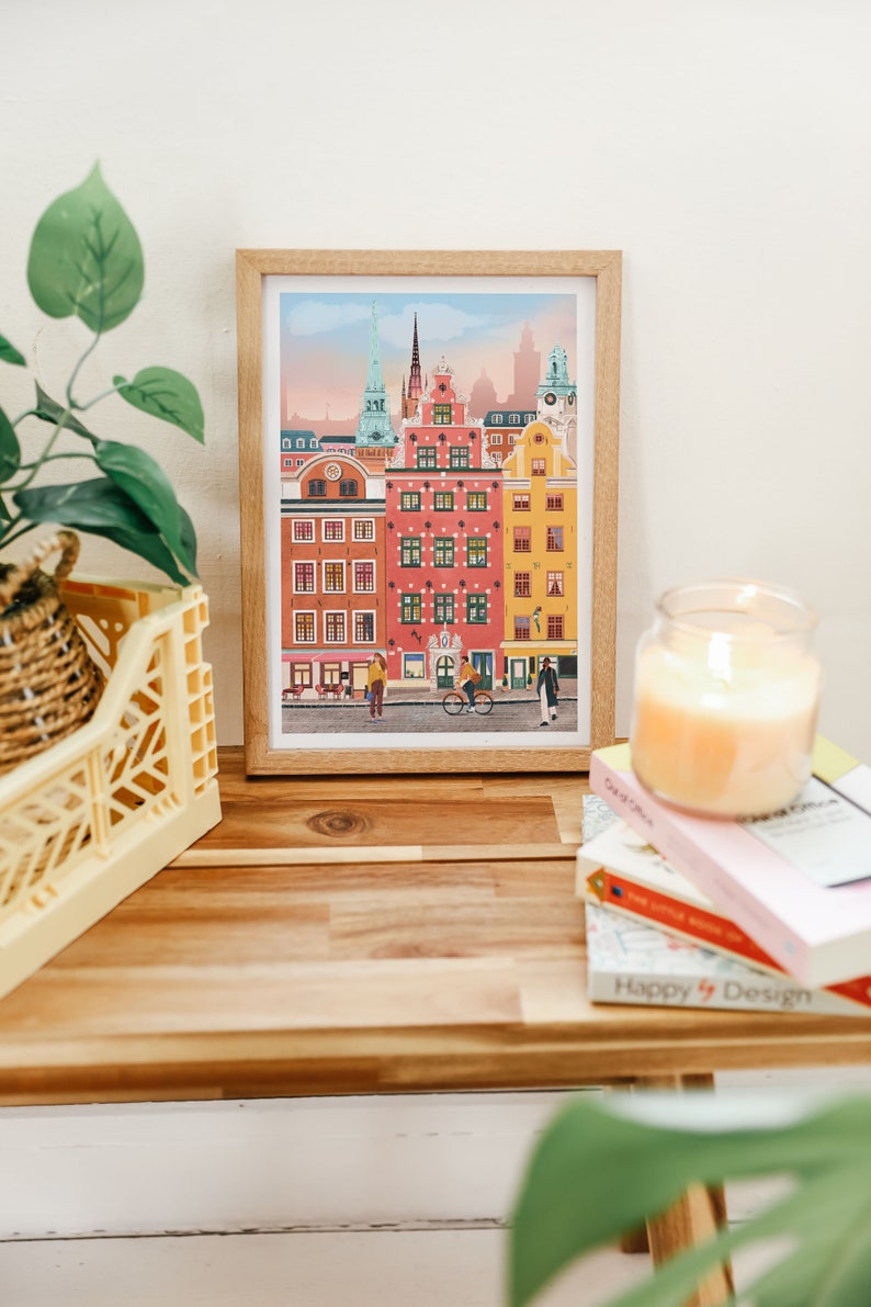 Pode incluir: Uma ilustra&ccedil;&atilde;o emoldurada de uma cena de rua da cidade com edif&iacute;cios coloridos, uma bicicleta e pessoas caminhando. Os edif&iacute;cios est&atilde;o em uma paleta de cores pastel com detalhes em rosa, amarelo e azul. A ilustra&ccedil;&atilde;o &eacute; emoldurada em uma moldura de madeira clara.