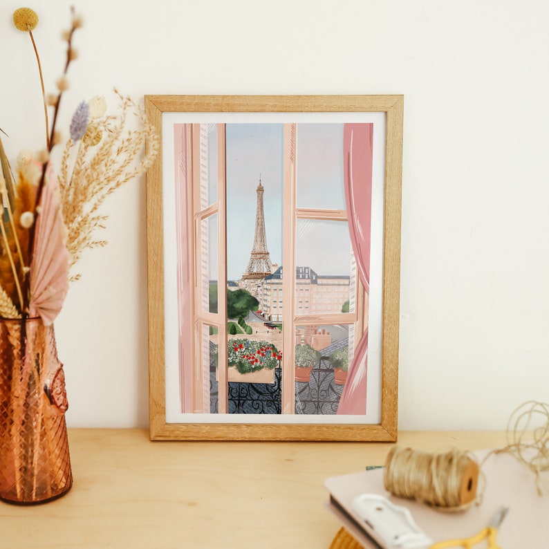 Op de afbeelding: Een ingelijste illustratie van een raam met uitzicht op de Eiffeltoren in de verte. Het raam is open en heeft roze gordijnen. Er staan bloemen op het balkon.