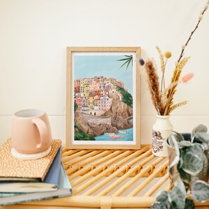 May include: A framed art print depicting a colorful coastal village scene. The artwork features buildings in various colors, set against a blue sky and sea. A pink mug, books, and decorative vase with dried flowers are also visible.